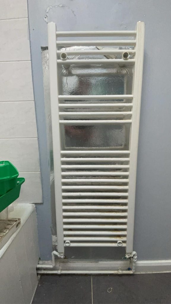 BEFORE: Old, worn-out white bathroom radiator with rust and peeling wall paint.