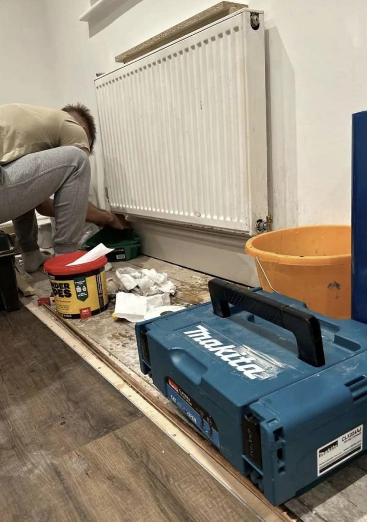 Technician fitting a new white radiator in a living room with tools and construction materials on the floor.