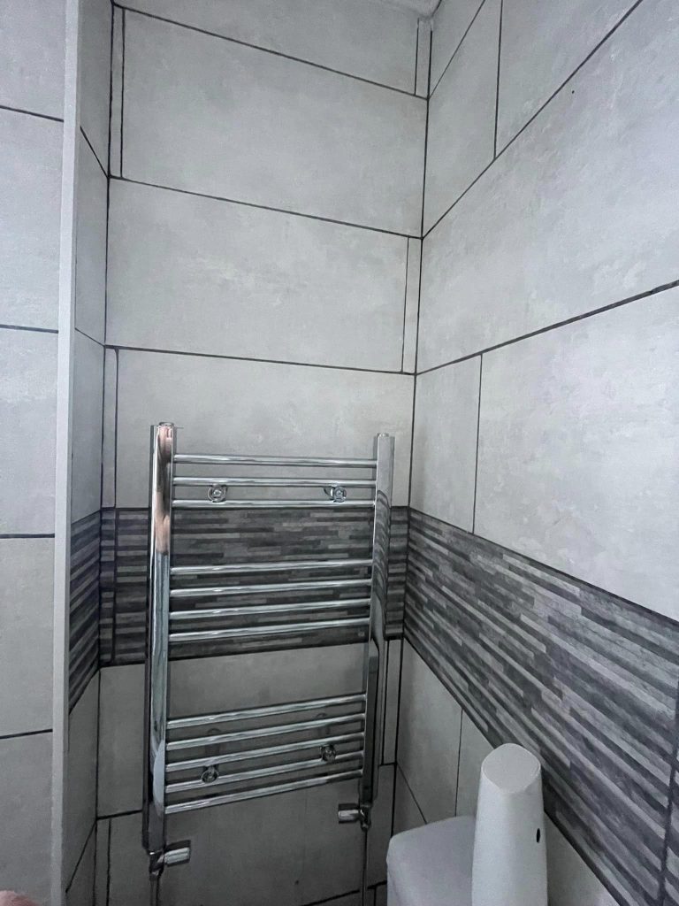 Perspective: This image provides a wider view of the chrome towel rail within the customer's bathroom, giving a sense of scale against the full height of the tiled wall.